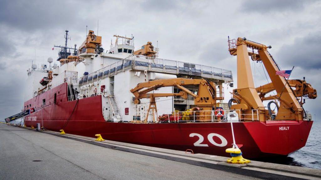 The image shows the US Coast Guard research ship, the Cutter Healy. It has a red hull and a yellow crane on the back. It has a large number 20 on the rear port side. 