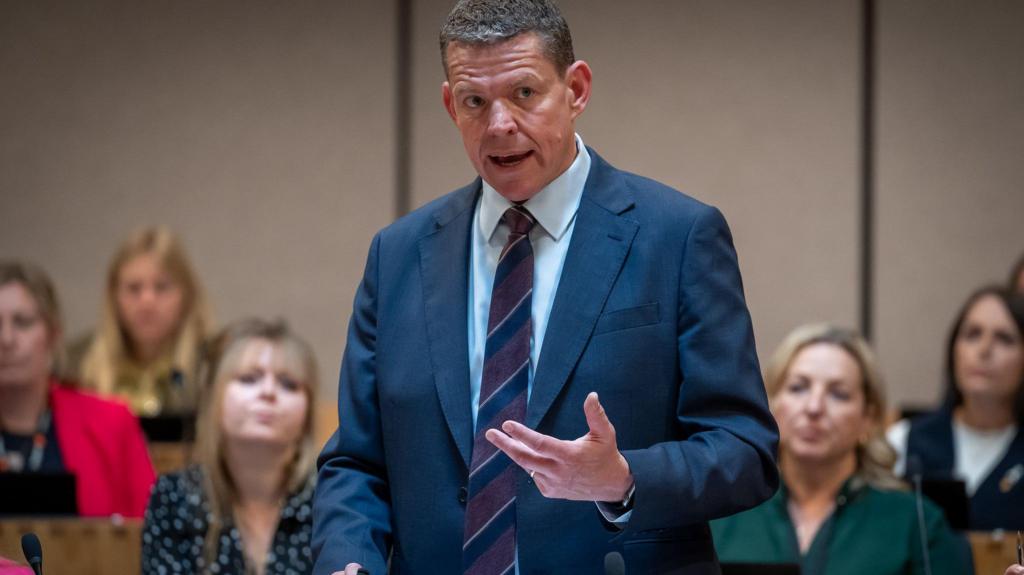 Rhun ap Iorwerth speaking in a suit and tie, in front of a crowd of his Senedd members who are sat behind him. He is stood up.