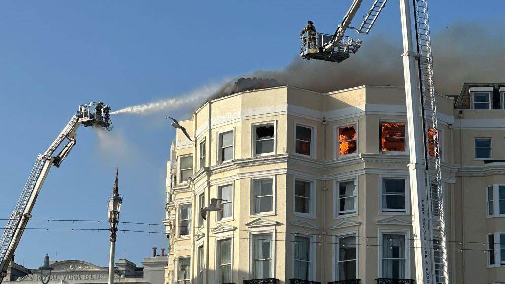 A shot showing two firefighters on raised aerial platforms firing water towards the fire that can be seen in a top-floor room.