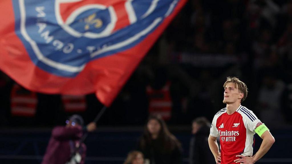 Arsenal captain Martin Odegaard looks disappointed as a large PSG flag is waved in the background.
