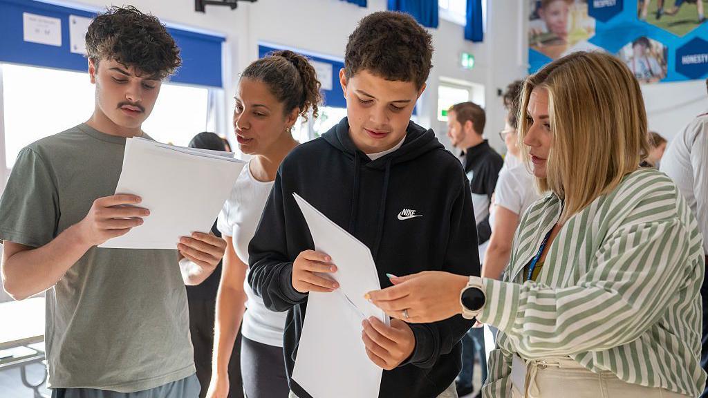 Two boys open their GCSE results as two women look at the results with them in a classroom.
