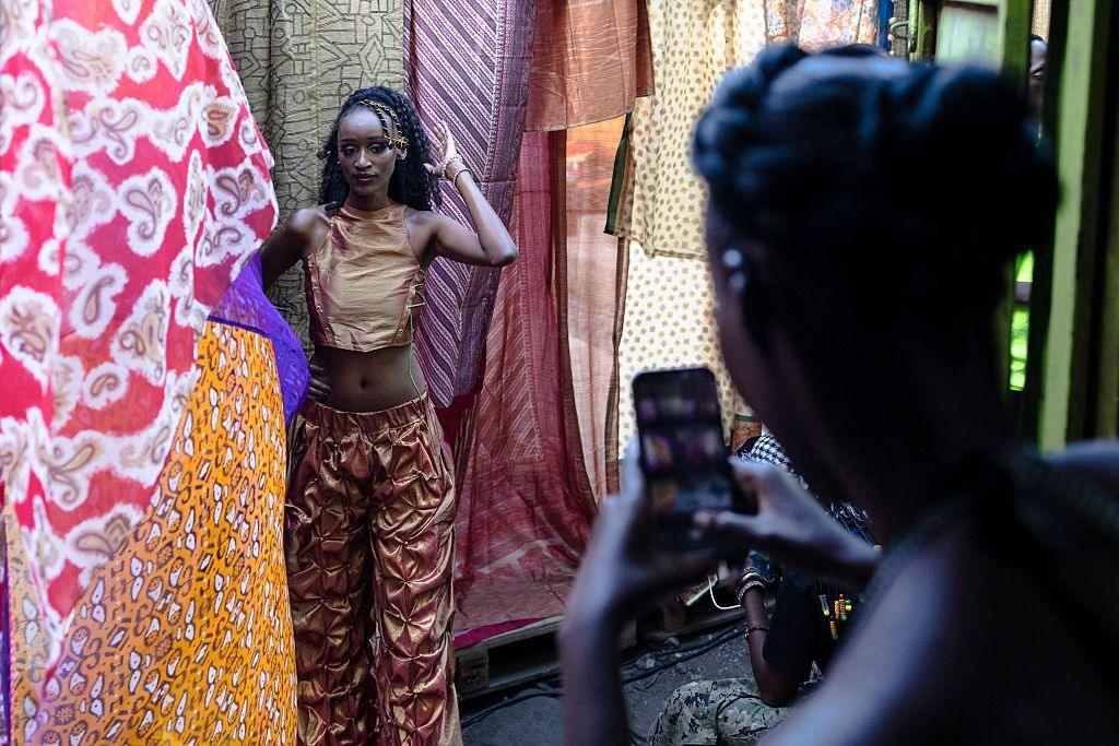 A woman wearing a bronze-coloured top and trousers stands next to draped pieces of colourful fabric while having her photo taken.