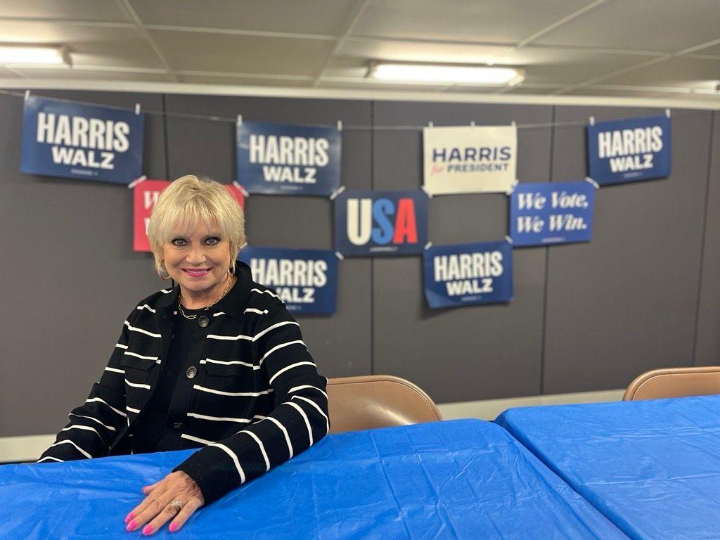 Lori McFarland poses in front of Harris Walz signs while wearing a black and white striped sweater