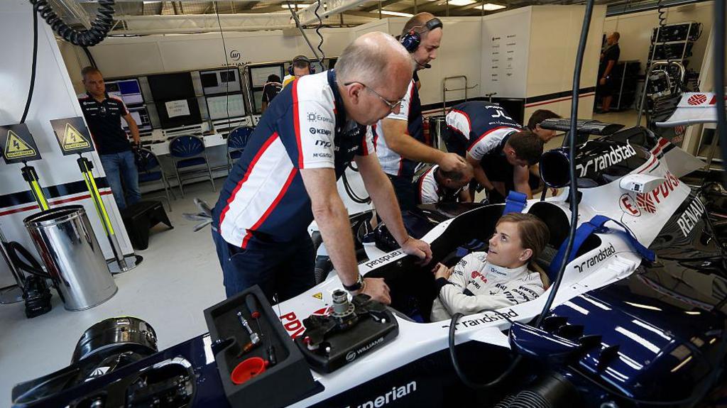 Susie Wolff in white driving suit is sitting low in a blue and white Formula 1 car. It is in a garage and four men are working on the rear of the car. Another man is standing over her, talking. All the men are wearing the blue, red and white Williams team colours.
