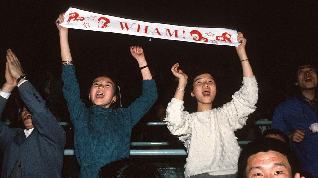 Audience members cheer during a Wham! concert, with two fans holding a white scarf, bearing the band's name and images in red, above their heads. 