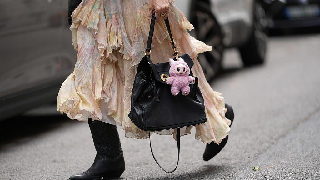 A pink Labubu doll clipped to a black handbag, carred by a woman