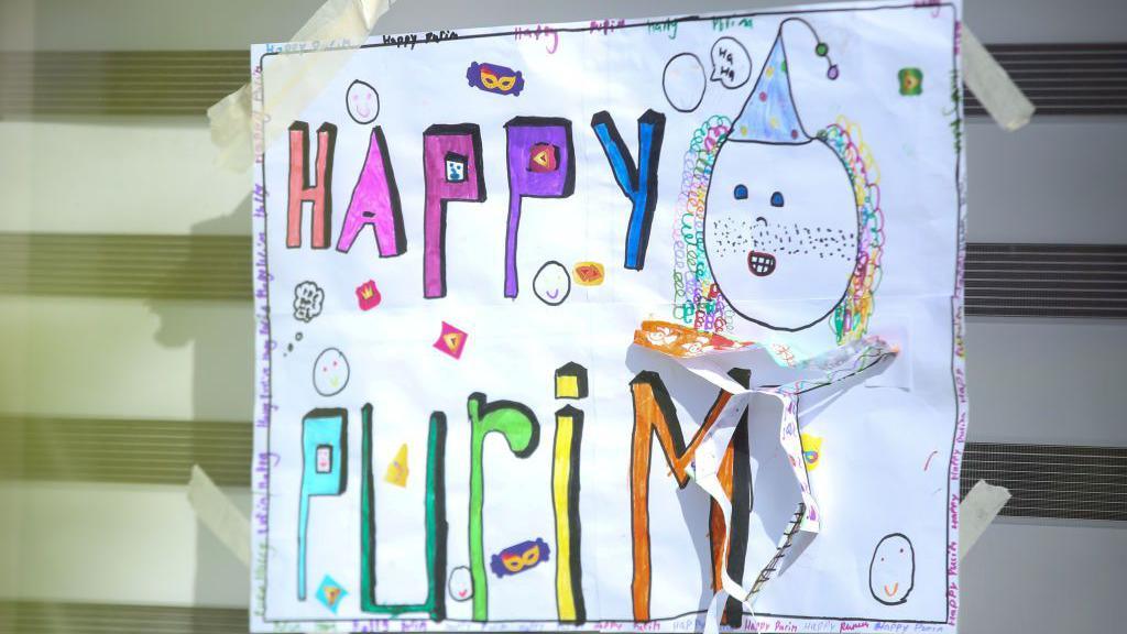 A child's poster saying "Happy Purim" in colourful writing is stuck on a window. 