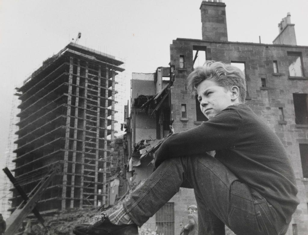 A black and white image of a young Gorbals boy sitting on a wall in front of a tenement in the middle of being demolished, with the bones of a tower block under construction beyond it.