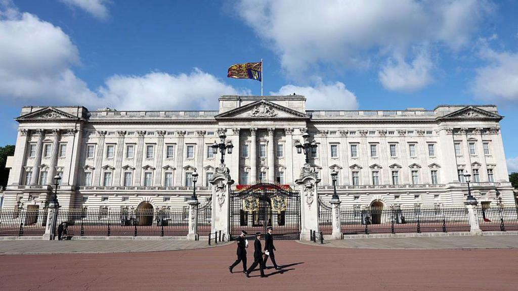 people walk past the front gate of buckingham palace, which has been dressed for trooping the colour. the royal standard flies above the palace, contrasting against a bright blue sky.