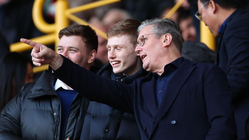 Keir Starmer at a football match in the stands pointing 