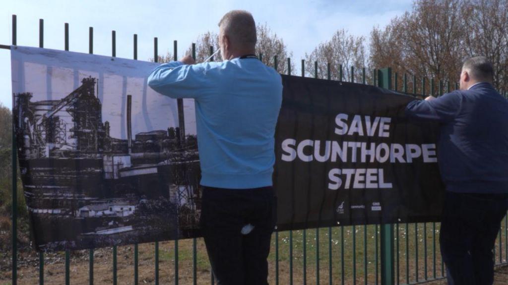 Two people are attaching a large banner to a green metal fence outdoors. The banner displays an image of a steel plant and reads 'SAVE SCUNTHORPE STEEL' in bold white letters. Trees and a clear sky are visible in the background.