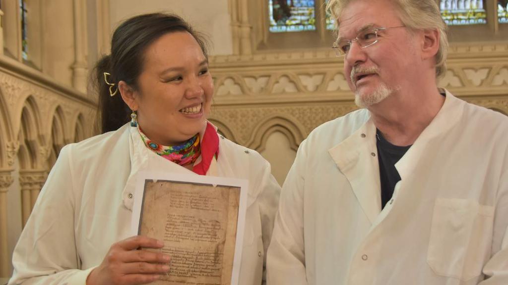 Dr Betty Chung with dark hair pulled back in a pony tail and a red neck scarf, holding a copy of the 9th Century manuscript page, and Prof John Carr with white hair and beard and wearing rectangle glasses, both wearing white lab coats in Corpus Christi College chapel