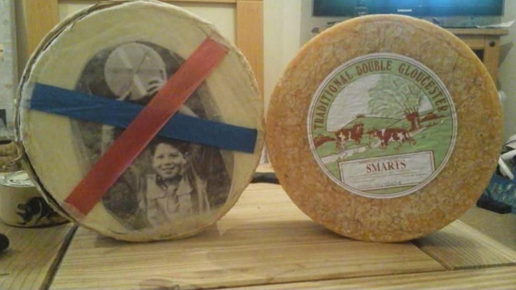 Two wheels of cheese used for cheese rolling sit on a wooden table. One bears the black and white image of a little boy holding a wheel of cheese, while the other has branding of traditional Double Gloucester cheese, with an image of cows grazing in a field.
