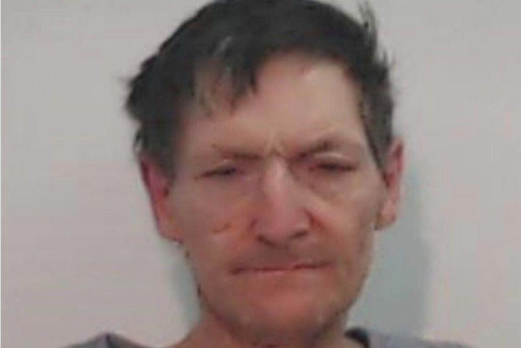 Blurry police mugshot of a dishevelled, expressionless man with his eyes barely open. He has a distinctive scar on his right cheek and unkempt dark hair. 