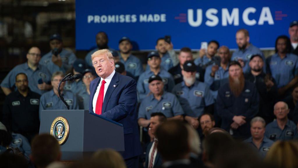 US President Donald Trump stands and speaks at a podium in front of auto workers with a "Promises Made USMCA" banner in the top right corner. 