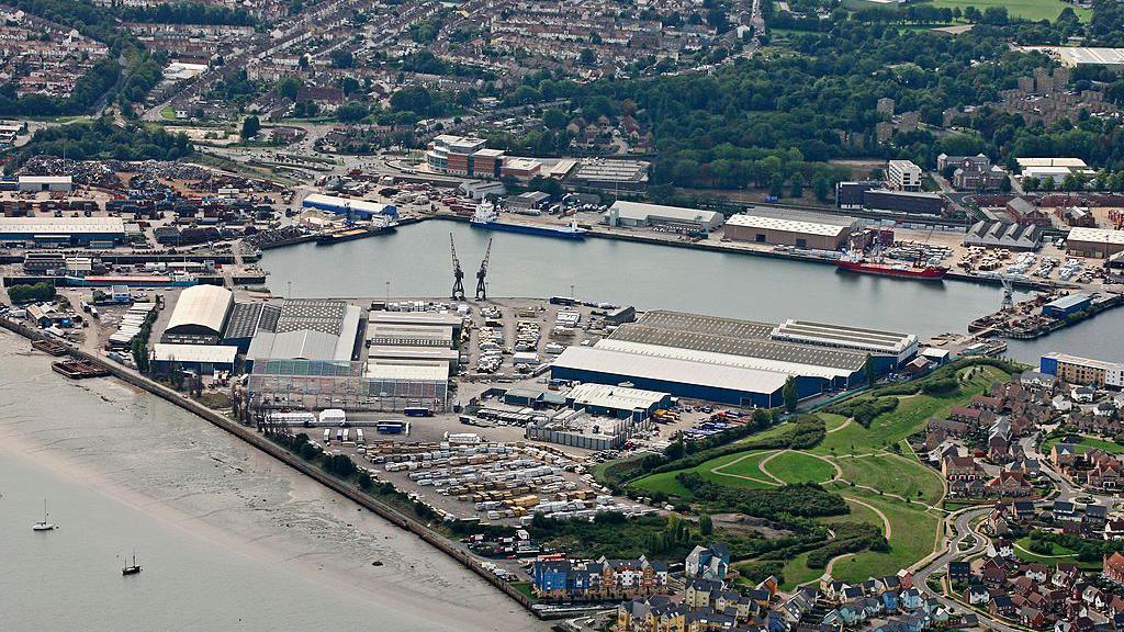 An aerial view of the Chatham Dockyards and surrounding areas. It is a block of water next to the coast and surrounded by hangar-type buidings.