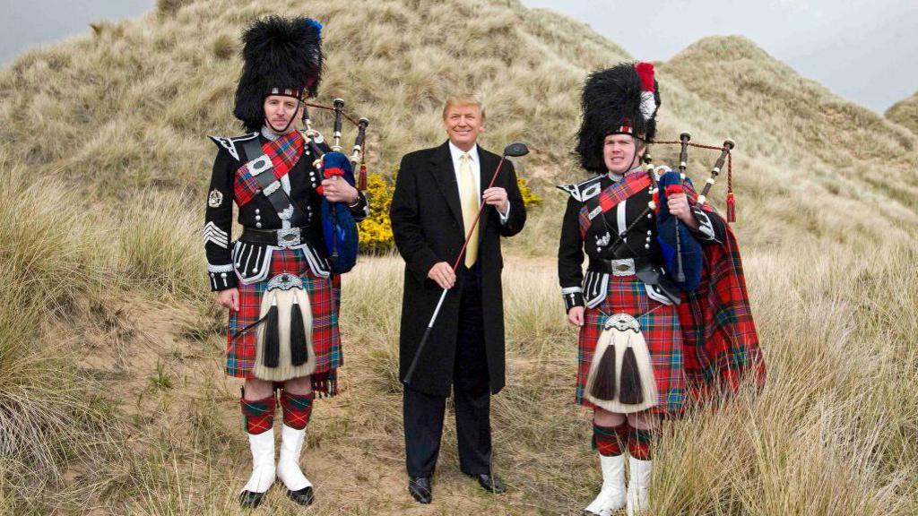 A photo from 2010 of Donald Trump in the sand dune at Menie holding a golf club. He is flanked by two bagpipers in full Scottish regalia, kilt, sporran and feathered hat