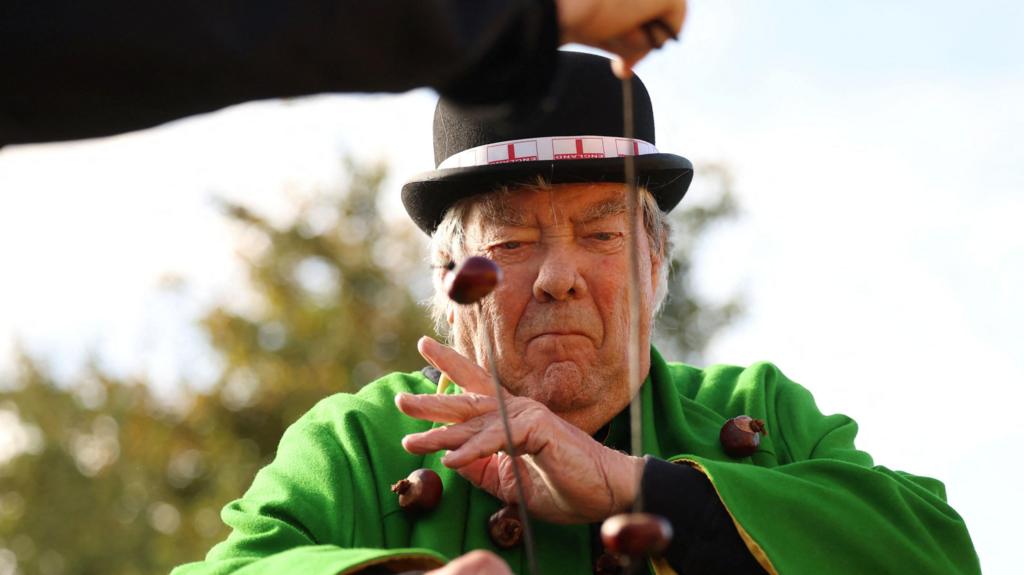 A man playing conkers. He is wearing a black bowler hat and bright green tunic. 
