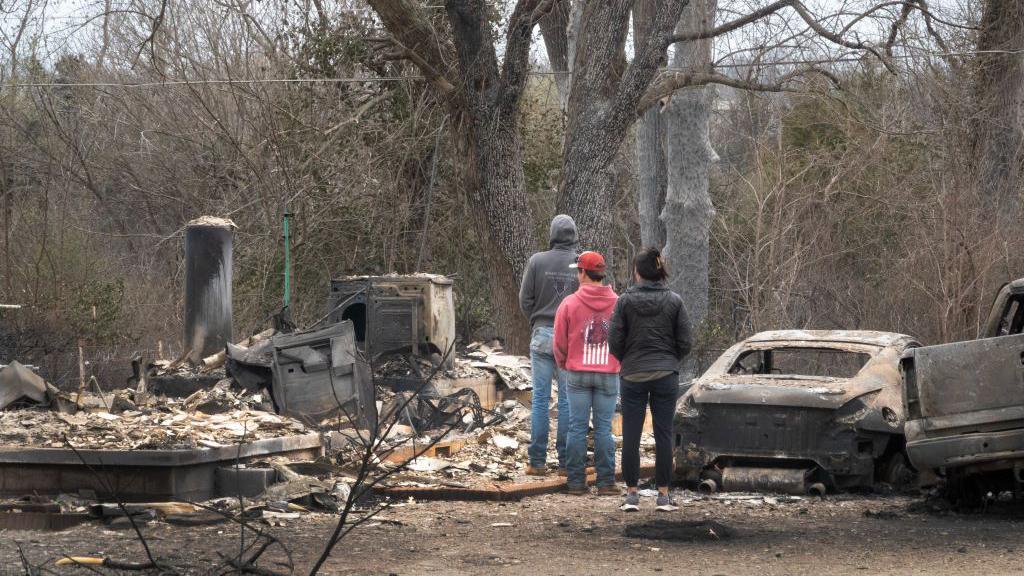 Three residents look at fire damage, which shows a house totally destroyed and a blackened car next to them.