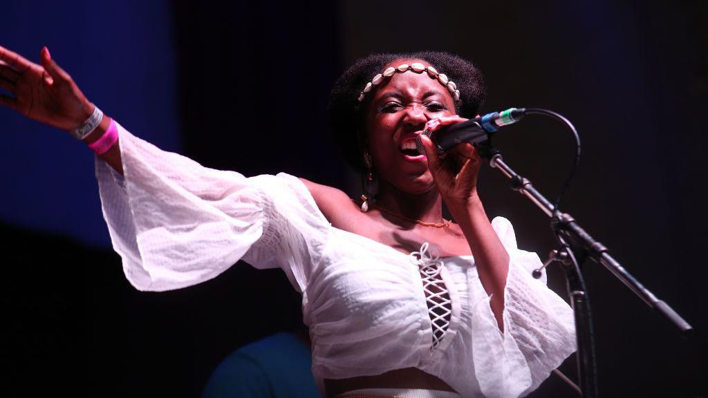 Nadine Caesar of Soul II Soul performs during Weyfest Festival 2021wearing a white shirt and singing in to a microphone with one hand in the air