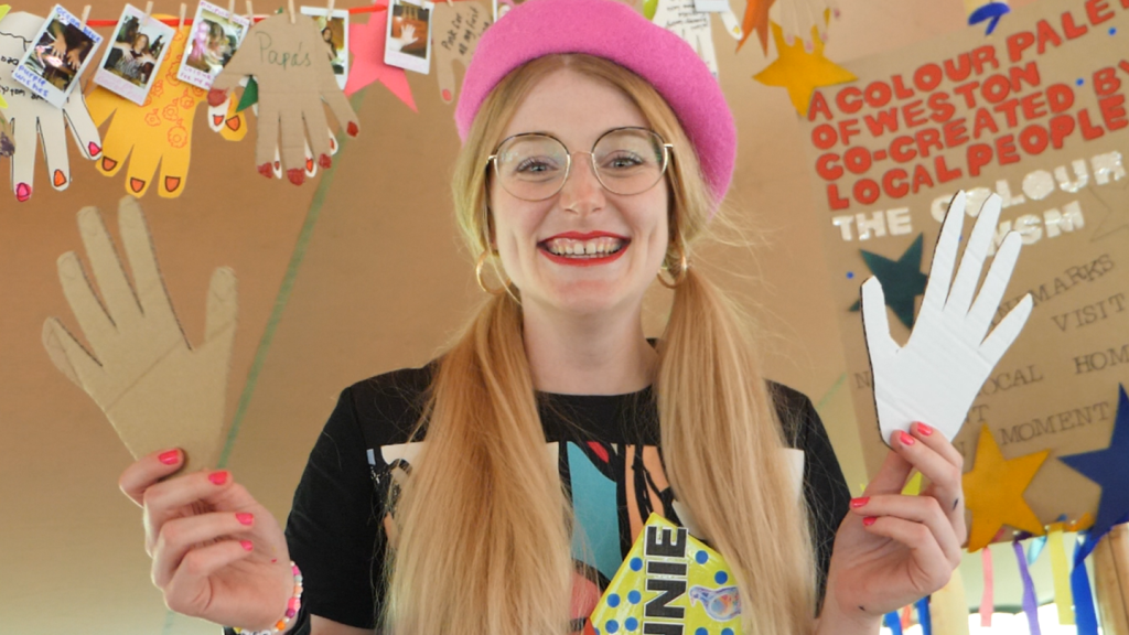 A person with their blonde hair in bunches wearing a pink beret , glasses and red lipstick, holding two cardboard hands