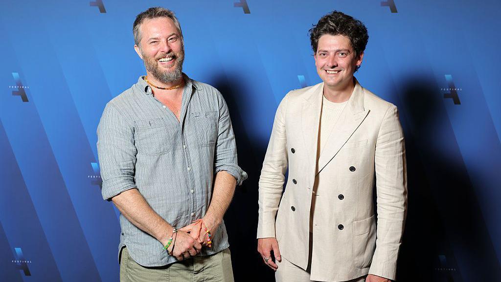 Two men stood in front of a blue background, smiling and looking at the camera. The man on the left has grey hair and a beard and is wearing a light grey linen shirt and a beaded brown necklace. The man on the right has curly dark hair and is wearing a cream t shirt and suit jacket.
