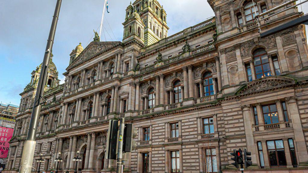 Glasgow City Chambers on George Square with pedestrians walking by and a lamp post and traffic lights in the foreground. 