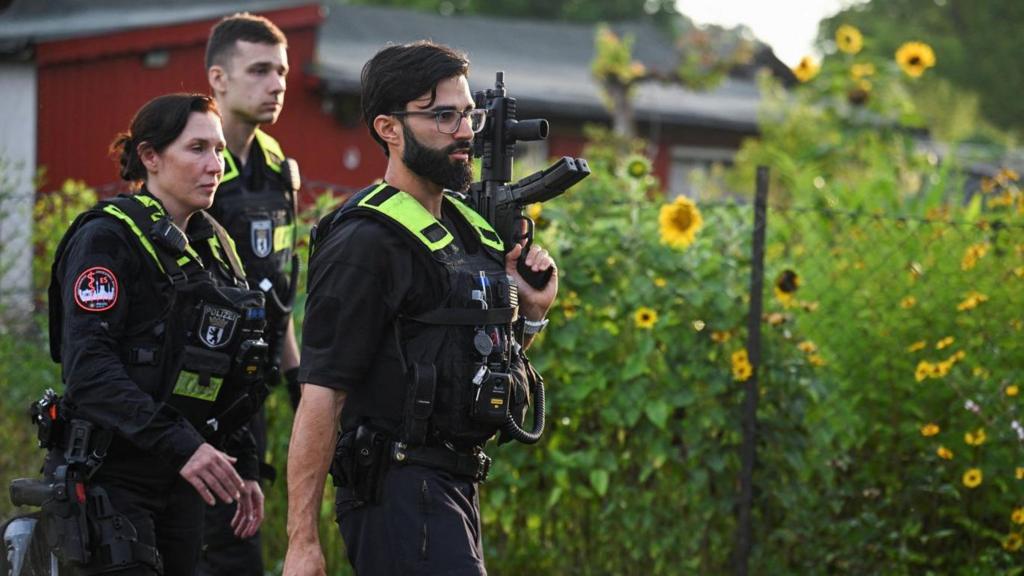 Police officers with firearms are shown walking in front of a fenced garden.