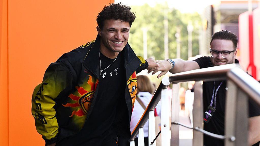 Lando Norris smiling while wearing a black and yellow jacket with a sun motif