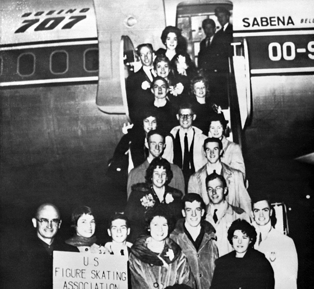 People stand outside a plane holding a sign that reads "US Figure Skating Association" in black and white photo