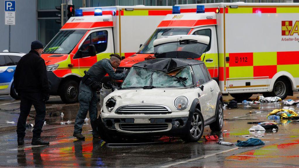 Two emergency service workers inspect a damaged white mini cooper.