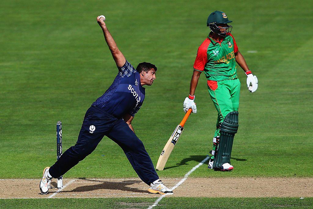 Majid Haq of Scotland bowls during the 2015 ICC Cricket World Cup match between Bangladesh and Scotland at Saxton Field on March 5, 2015 in Nelson, New Zealand. He is side on, wearing blue and bowling full pelt at the other wicket from left to right of shot. A Bengladeshi player in green and red is standing behind him at the other wicket.