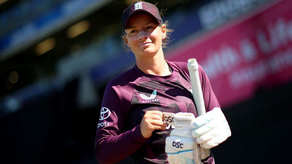 Danni Wyatt-Hodge holding her cricket bat and gloves while wearing England's purple training kit and cap