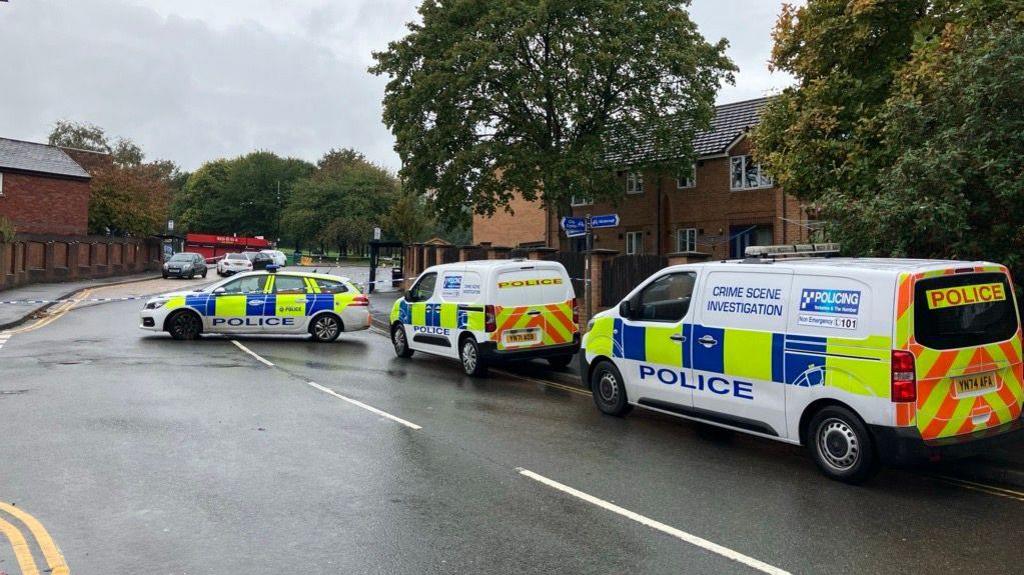 The image depicts a street that has been cordoned off by police, with three marked police vehicles blocking access. These vehicles feature high-visibility livery and include signage indicating "POLICE" and "CRIME SCENE INVESTIGATION". The surrounding area includes residential buildings and trees. The sky is overcast, and the wet road surface suggests that it has recently rained.