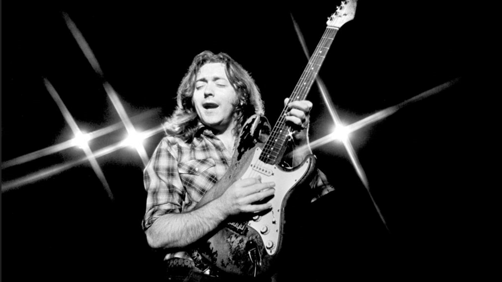 Rory Gallagher, with long hair and checked shirt, playing a guitar