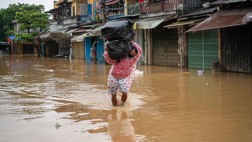 A man in a raincoat wades through the floodwaters in Hoi An, carrying two large packs of items wrapped in black trash bags