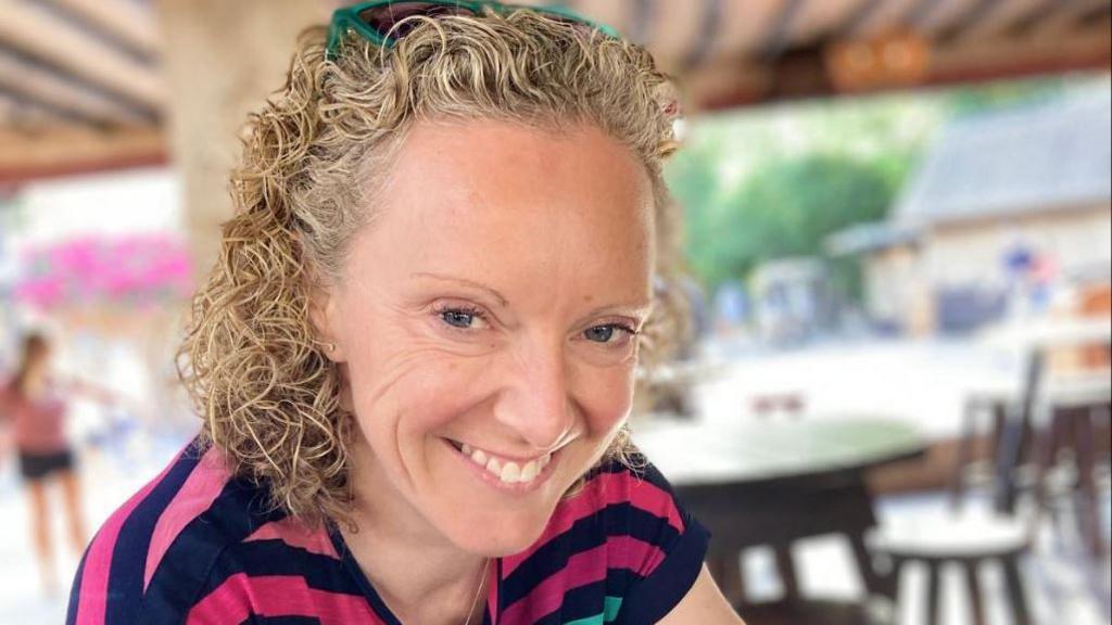 A woman with curly blonde hair smiles at the camera. She is wearing a stripy blue and pink T shirt.