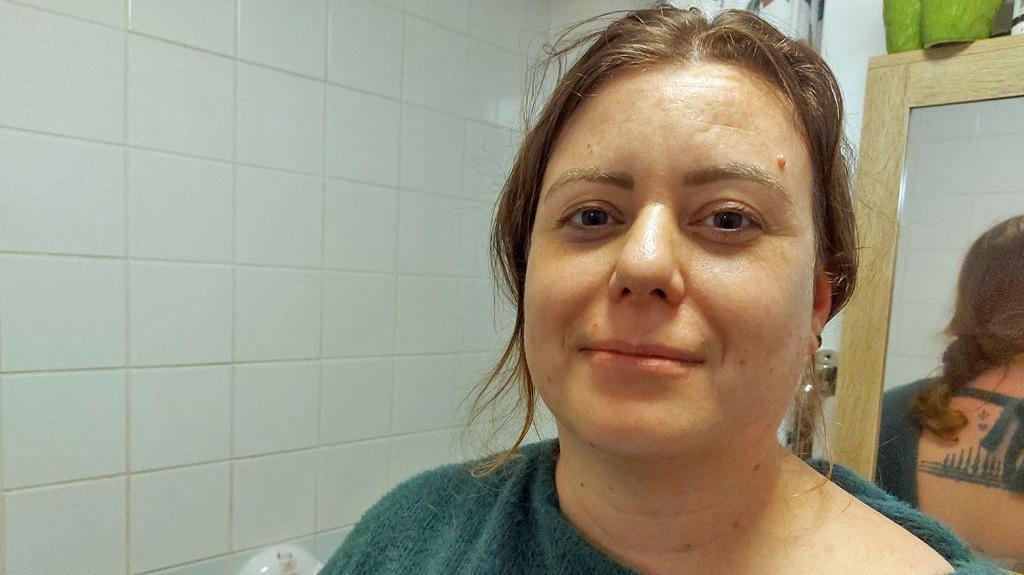 Headshot of Sam in her bathroom. She has wavy brown hair tied back, with some strands framing her face. She is wearing a green off-the shoulder jumper - and reflected in the mirror behind her you can see it is tied at the back and she has a tattoo. The wall behind her has white tiles, there's a shower curtain and wooden framed bathroom cabinet. 