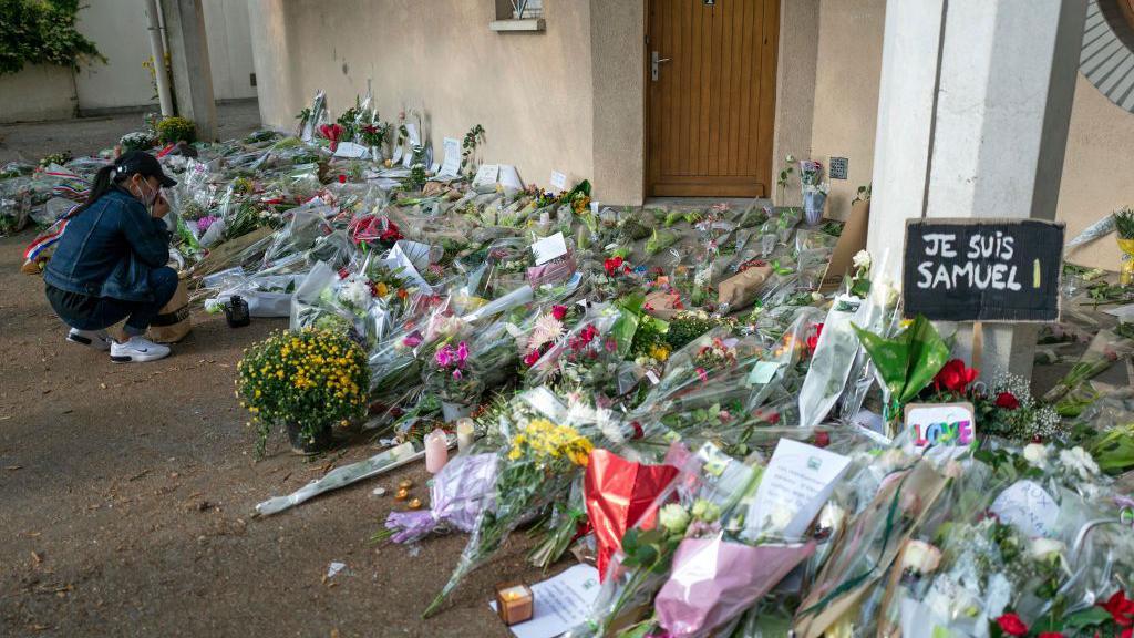 A woman crouches down to look at dozens of bunches of flowers left on the ground near a building. A sign reads "Je suis Samuel"