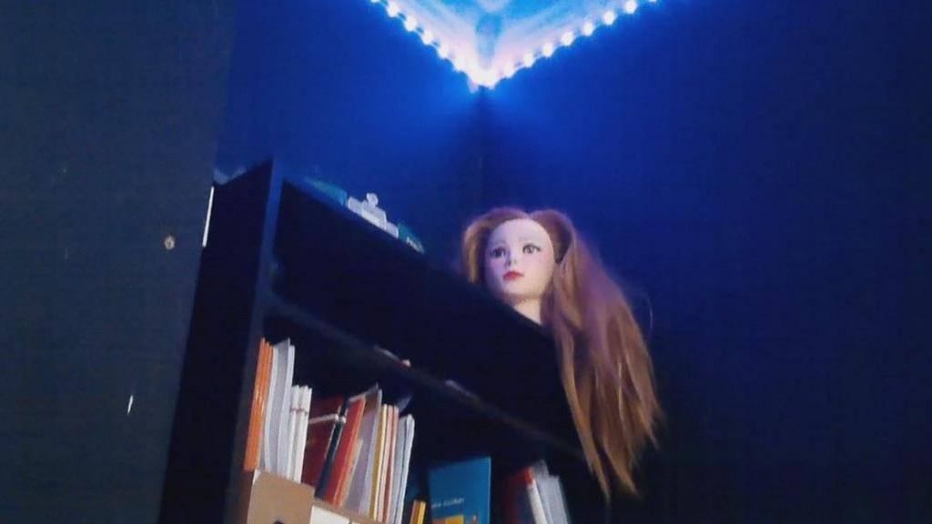 A doll's head with long red hair sits on top of a black bookshelf inside the school's sensory room. A bright fluorescent light shines above it. The surrounding walls are all black.