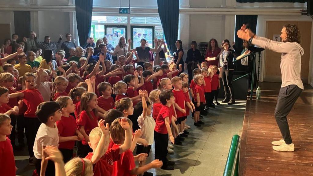 Image of a room filled with young schoolchildren, wearing red tops and black shorts and trousers. They are all looking at a stage, with their arms in the air, copying a man with curly hair who is wearing a grey hoodie, black trousers and white trainers. In the background can be seen a number of adults standing against a large window watching.