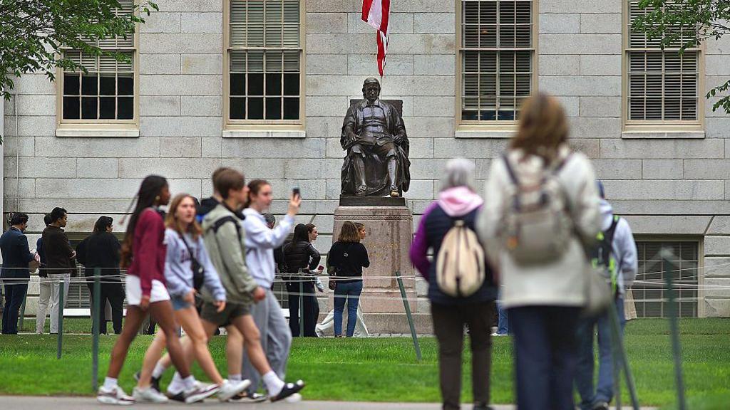 Tour groups walk past the statue of John Harvard in Harvard Yard on May 2, 2025. The photo shows a group of students with backpacks also walking past the statue.