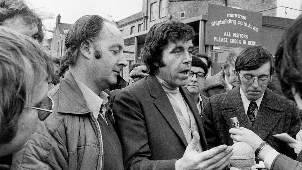 A black and white shot of a man speaking to journalists - who are out of the shot. He is surrounded by other men and is standing in front of a sign which reads "Marathon shipbuilding co"