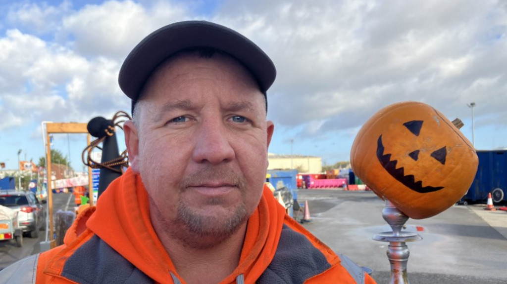 man wearing orange hooded jacket wearing baseball cap standing next to a display of a pumpkin head on a candelabra at a tip, there's a cloudy sky.
