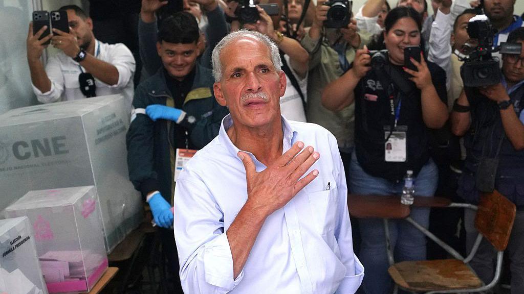 An image showing Tito Asfura at a polling station in Honduras. Behind him are spectators who have their phones out to capture the moment. Asfura is in the centre of the image, wearing a white button-up shirt rolled at the sleeves. He is gesturing with his right hand towards his chest. 