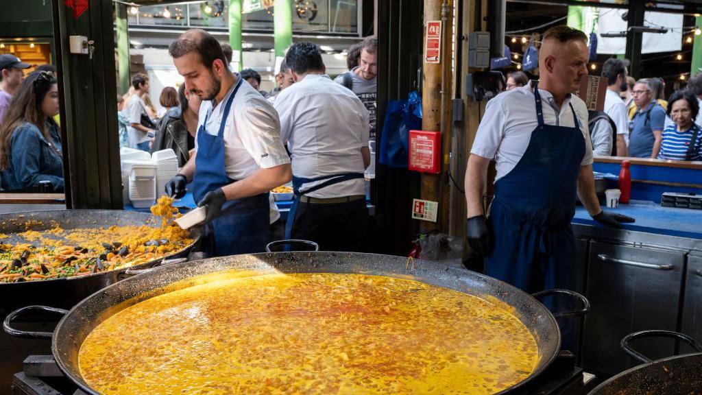 Large pans of street food paella at Borough Market. A giant cooking vessel is filled with yellow rice and liquid and men wearing aprons and gloves stir and serve it