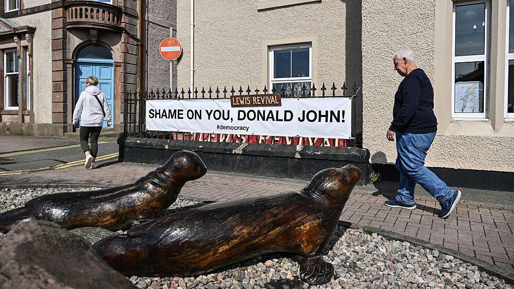 A street in Stornoway. Outside a cream-coloured harled building, a banner saying 'Shame on you, Donald John' is attached to railings. A woman and man are walking past, one behind the other. There are two bronze sculptures of seals on the ground in front of them.