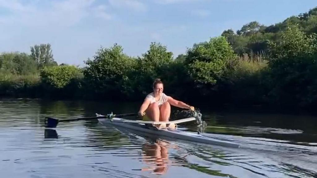 A young women with brown hair wearing a white top rowing on a thin rowing boat. She is mid stroke and sitting on a dark green river with trees lining the bank.