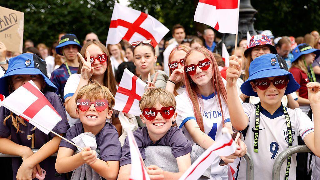 Six children standing at the front of the crowd holding England flags and wearing sunglasses with the names of Chloe Kelly and Grace Clinton written on them with pen marker.