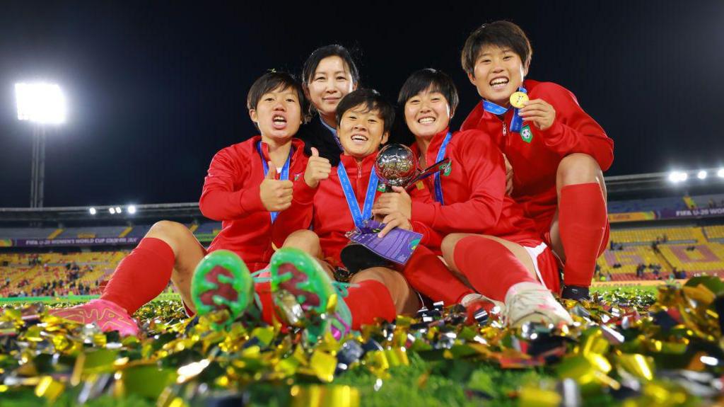 North Korea players celebrate and show off their medals to the camera. They're sitting on the turf, which covered in gold-colured tickertape.